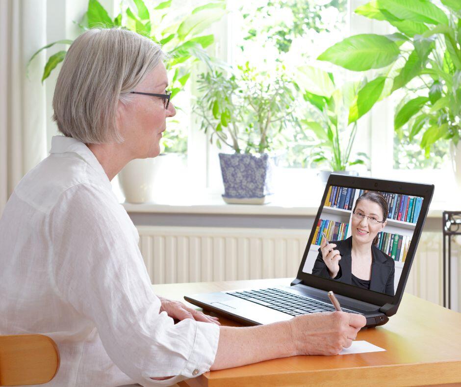 Seitliche Perspektive; ältere Frau mit Brille und weißer Bluse sitzt am Tisch auf dem ein Laptop mit einer Frau auf dem Bildschirm, die einen Vortrag hält