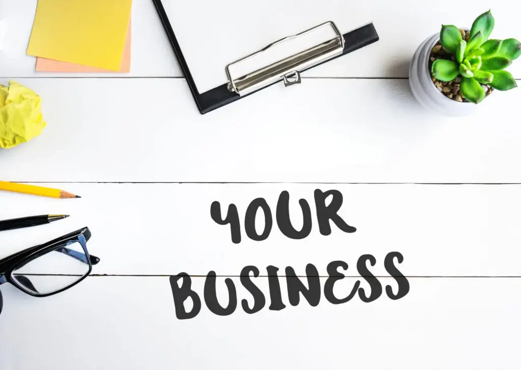 Draufsicht auf einen weißen Tisch mit Brille, Stiften, Pflanze und Klemmbrett und Text "Your Business"