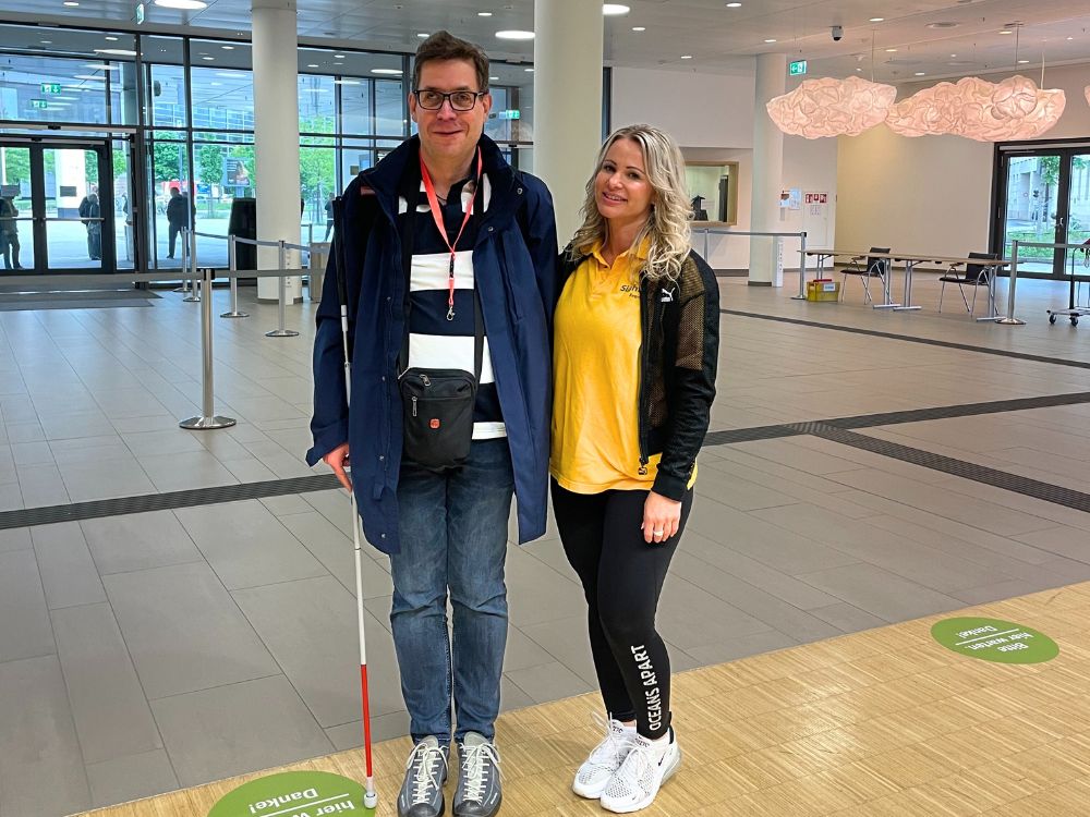 Blind man with white cane smiles at the camera together with blonde woman in yellow shirt with SightCity logo