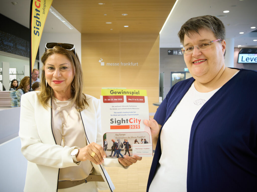 Two women stand side by side in a trade fair building, jointly holding a SightCity 2025 catalogue with a competition sticker attached. On the left, a woman with long brown hair and a white blazer; on the right, a woman with short hair and glasses wearing a dark blue jacket. In the background, the SightCity Infopoint banner and the 'messe frankfurt' lettering. Text on the catalogue: 'Gewinnspiel, 21 to 23 May 2025, SightCity 2025, Kap Europa, Congress Centre of Messe Frankfurt'.