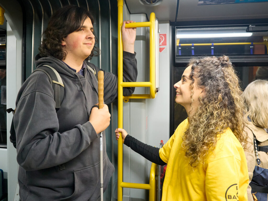 A young man with dark curly hair and a grey hoodie is standing inside a bus or tram, holding a white cane. He is smiling at a young woman wearing a yellow T-shirt with curly hair, who is holding onto a yellow handrail. Other passengers are visible in the background. The woman's yellow T-shirt bears a logo suggesting a guiding or orientation service.