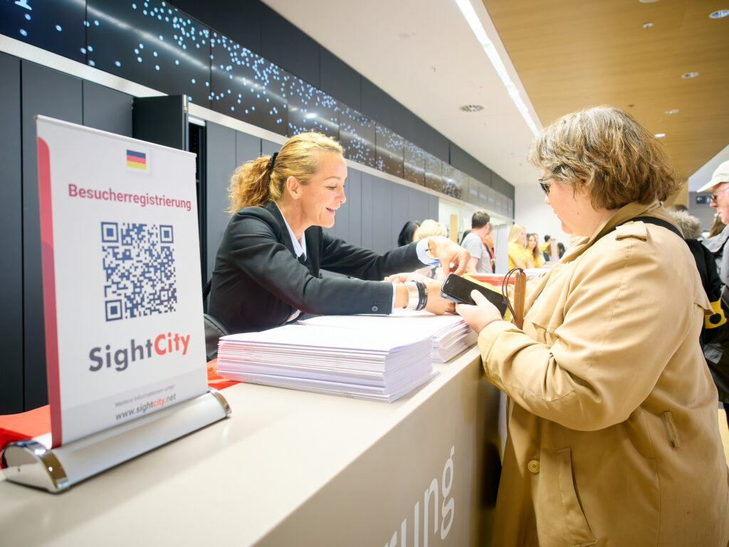 A staff member behind the SightCity registration desk is smiling and handing a ticket or badge to a visitor. The visitor is wearing a beige coat and dark sunglasses. A stack of programme booklets is on the counter. In the foreground, a display stand features the SightCity logo, a QR code, the text "Besucherregistrierung" (visitor registration), the German flag, and the web address www.sightcity.net. In the background, additional visitors and a decorative light installation are visible.