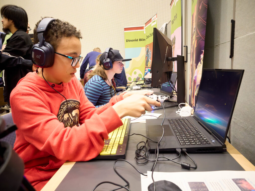 Two young people are sitting side by side at a table playing video games. The young person in the foreground is wearing a red sweater, glasses, and large headphones, operating a controller and a yellow backlit keyboard. A game is visible on the laptop screen in front of them. In the background, another child wearing headphones and a cap is also playing a game. A banner in the background reads "Dinonid Blin." The scene depicts an inclusive gaming activity.