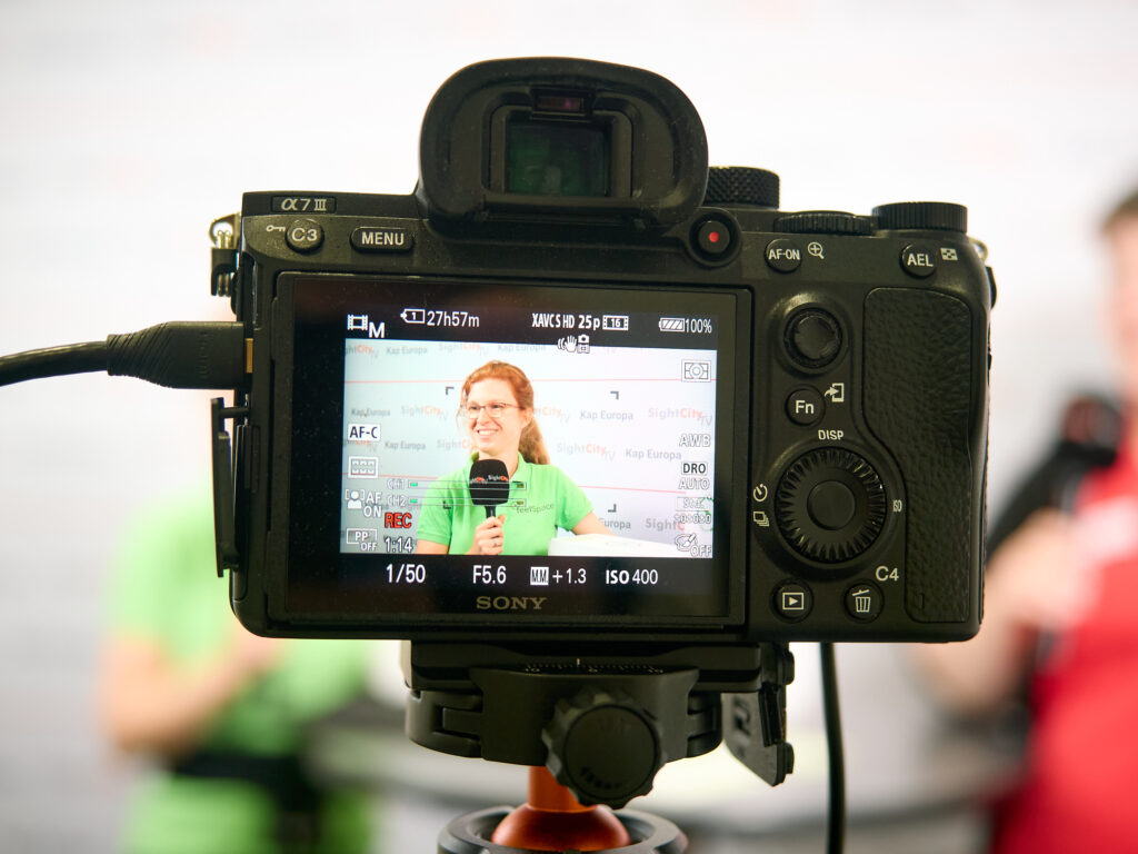 A view of the screen of a Sony Alpha 7 III camera mounted on a tripod. The display shows a woman with red hair, glasses, and a green SightCity polo shirt holding a microphone. In the background of the camera screen, a press wall with the labels "SightCity TV" and "Kap Europa" is visible. The camera is in recording mode (REC).