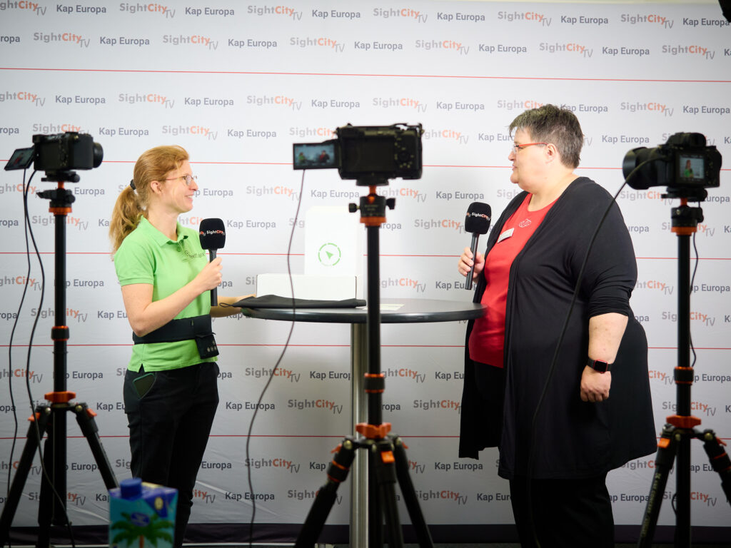 Two women are facing each other conducting an interview as part of SightCity TV. The woman on the left is wearing a green SightCity polo shirt and holding a microphone with SightCity branding. The woman on the right is wearing a red T-shirt under a black cardigan and is also holding a SightCity microphone. Between them stands a round high table with a white product box on it. In the foreground and on the sides, three cameras on tripods are visible recording the interview. In the background is a press wall with alternating text reading "SightCity TV" and "Kap Europa."