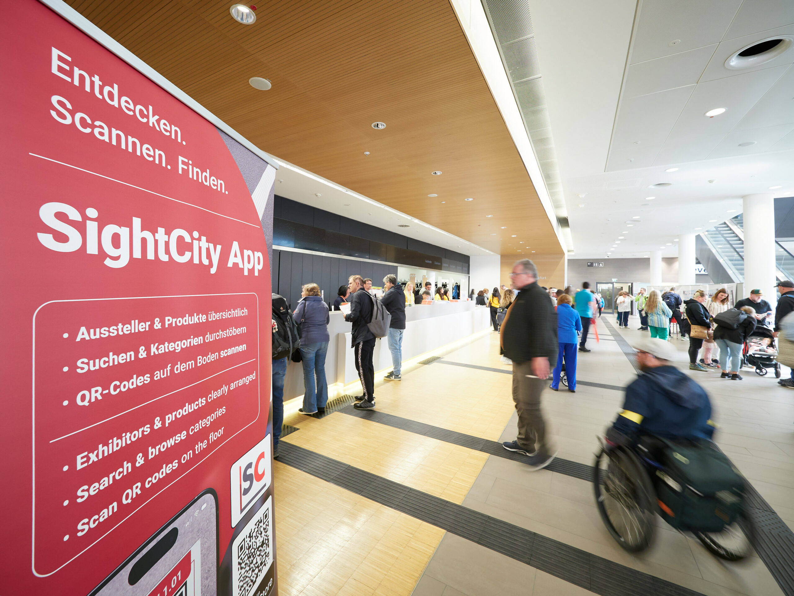 A view of the entrance area of SightCity. In the foreground, a large red roll-up banner reads "Entdecken. Scannen. Finden. – SightCity App" (Discover. Scan. Find. – SightCity App), listing the app's features in German and English: exhibitors and products clearly arranged, search and browse categories, scan QR codes on the floor. In the background, numerous visitors are moving through the bright entrance area. On the right side of the image, a person in a wheelchair is visible in motion.