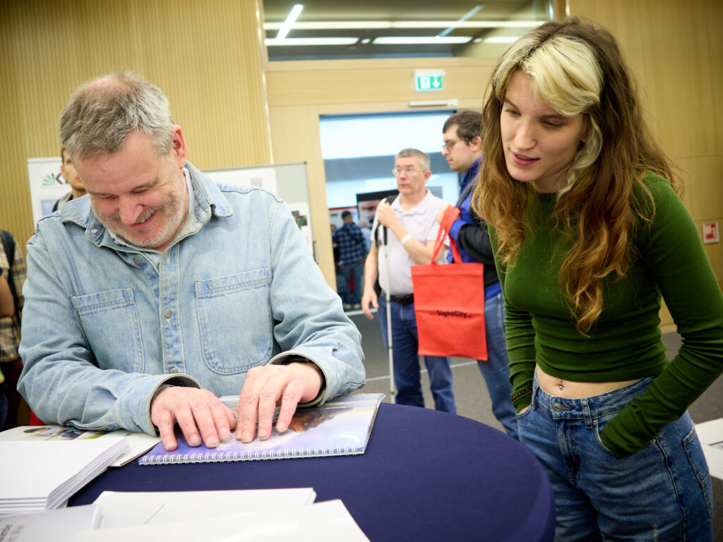 An older man with grey hair and beard sits smiling at a table, exploring an open document or brochure with both hands. Next to him stands a young woman with long brown hair and a green top, leaning towards him. In the background, other visitors and a man with a red SightCity bag and white cane. Scene from the SightCity trade fair.