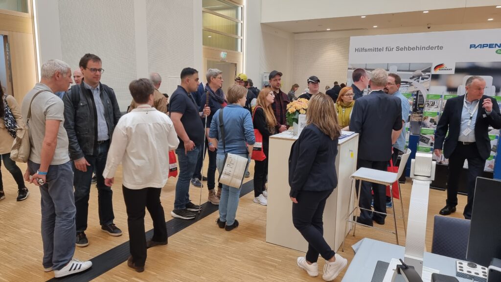 Busy trade fair aisle with numerous visitors, including several people using white canes. In the background on the right, the Papenmeier RehaTechnik exhibition stand with the banner 'Hilfsmittel für Sehbehinderte' (Aids for the visually impaired) and the Germany logo. Several people stand at the booth receiving consultations. The scene shows the lively atmosphere at the SightCity trade fair.