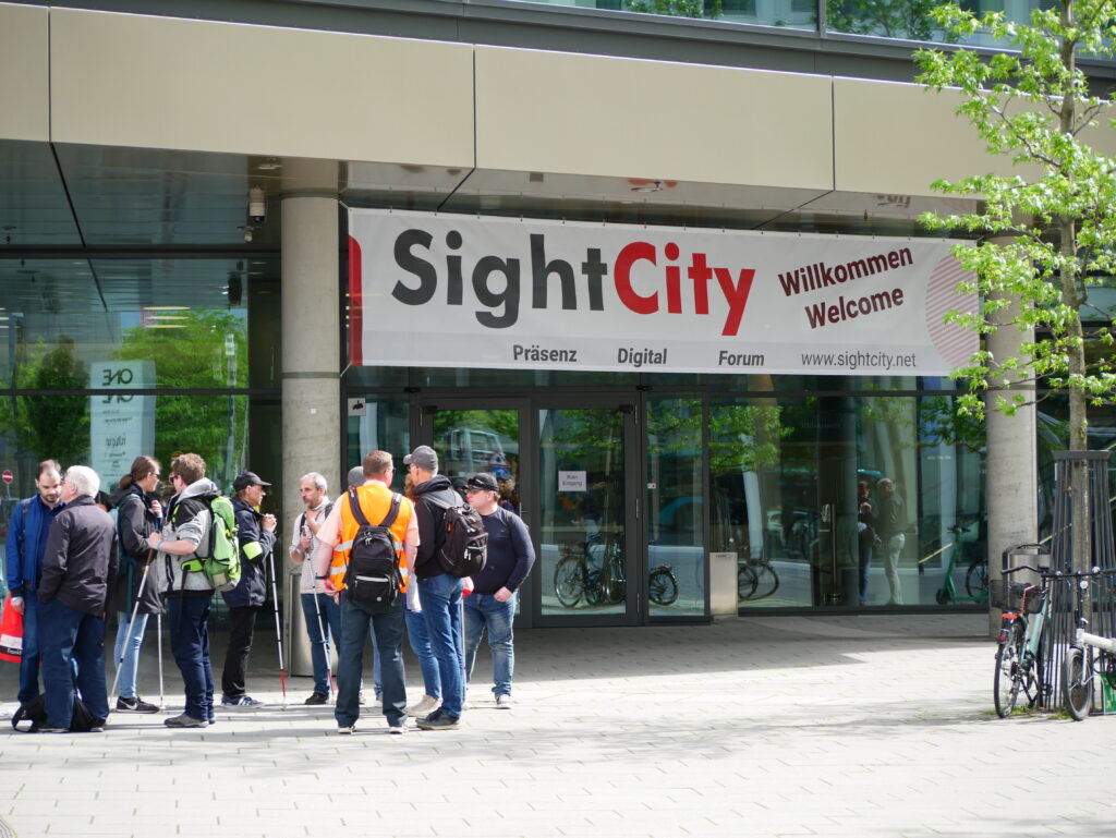 Exterior view of the SightCity entrance area. Above the glass doors hangs a large white banner reading: 'SightCity – Willkommen / Welcome – Präsenz – Digital – Forum – www.sightcity.net'. Several people stand in a group in front of the entrance, some with white canes. Sunny weather, trees in the background.