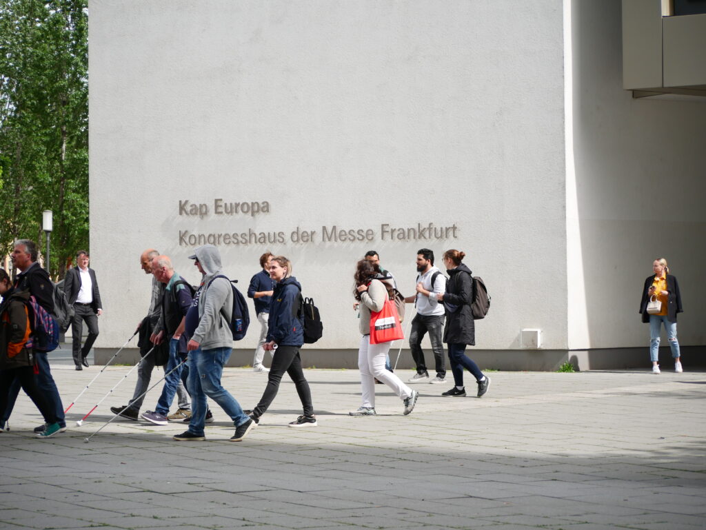 Exterior view of Kap Europa, Congress Centre of Messe Frankfurt. The inscription 'Kap Europa – Kongresshaus der Messe Frankfurt' is visible on the light-coloured facade. Numerous people walk across the forecourt, several with white canes and red SightCity tote bags. Sunny day with trees in the background.