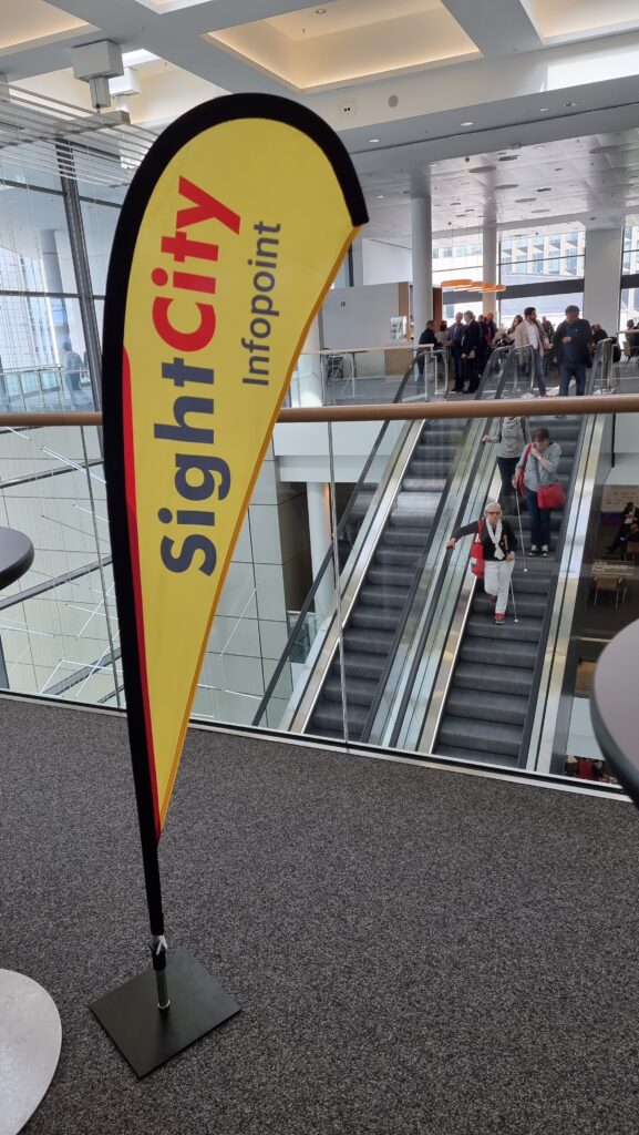 Yellow teardrop-shaped beach flag reading 'SightCity Infopoint' in red and black, placed on a black base inside the event venue. In the background, an escalator with a person using a white cane, and further visitors on a gallery level.
