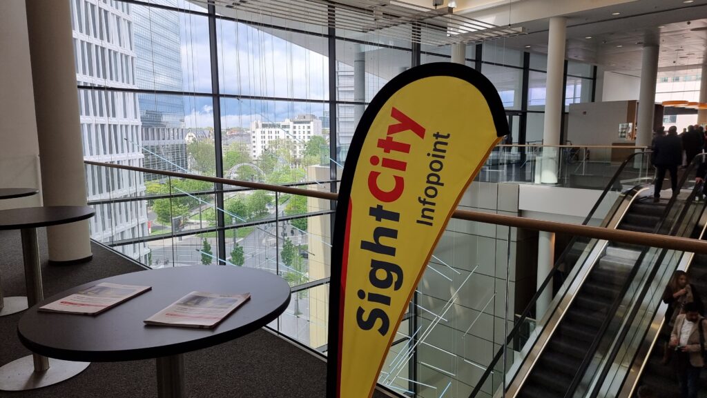 Yellow teardrop-shaped beach flag reading 'SightCity Infopoint' in red and black, placed next to a round standing table inside the event venue. Through the large glass front, the Frankfurt city skyline with high-rise buildings and green areas is visible. To the right, an escalator with visitors.