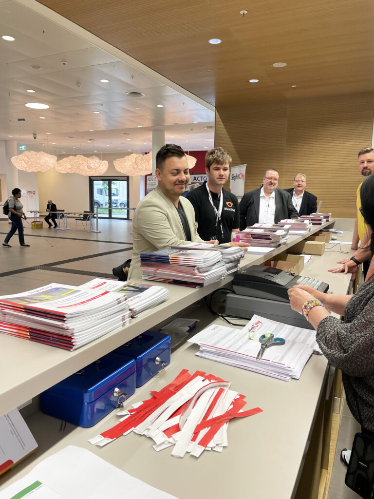 View of the SightCity registration desk. Several people stand behind the long counter, including two young men in the foreground – one in a light blazer, one in a black BlindShell-logo T-shirt. On the counter are stacked catalogues, red-and-white accreditation lanyards, cash boxes, and documents. SightCity banners and exhibition areas are visible in the background. A visitor stands at the counter.