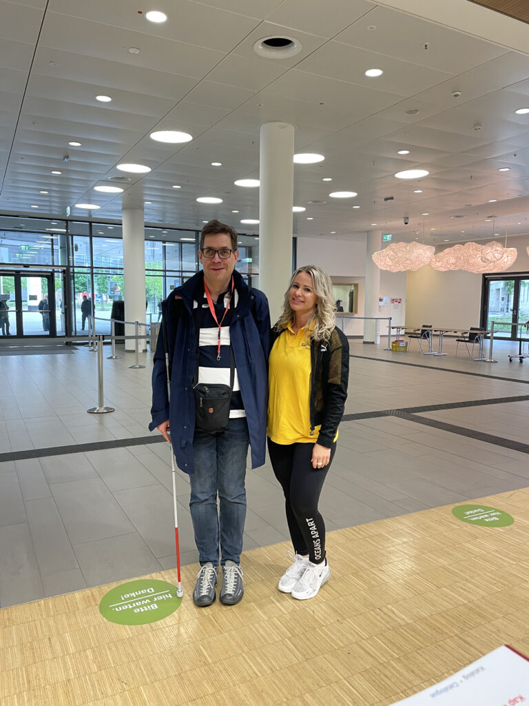 Two people stand side by side in the bright foyer of Kap Europa. On the left, a man wearing glasses, a red accreditation lanyard, a dark jacket, and holding a white cane. On the right, a woman with blonde curly hair, a yellow polo shirt with SightCity print, and a black jacket. In the background, the spacious foyer with columns, ceiling lights, and green floor markers reading 'Bitte hier warten. Danke!' (Please wait here. Thank you!).