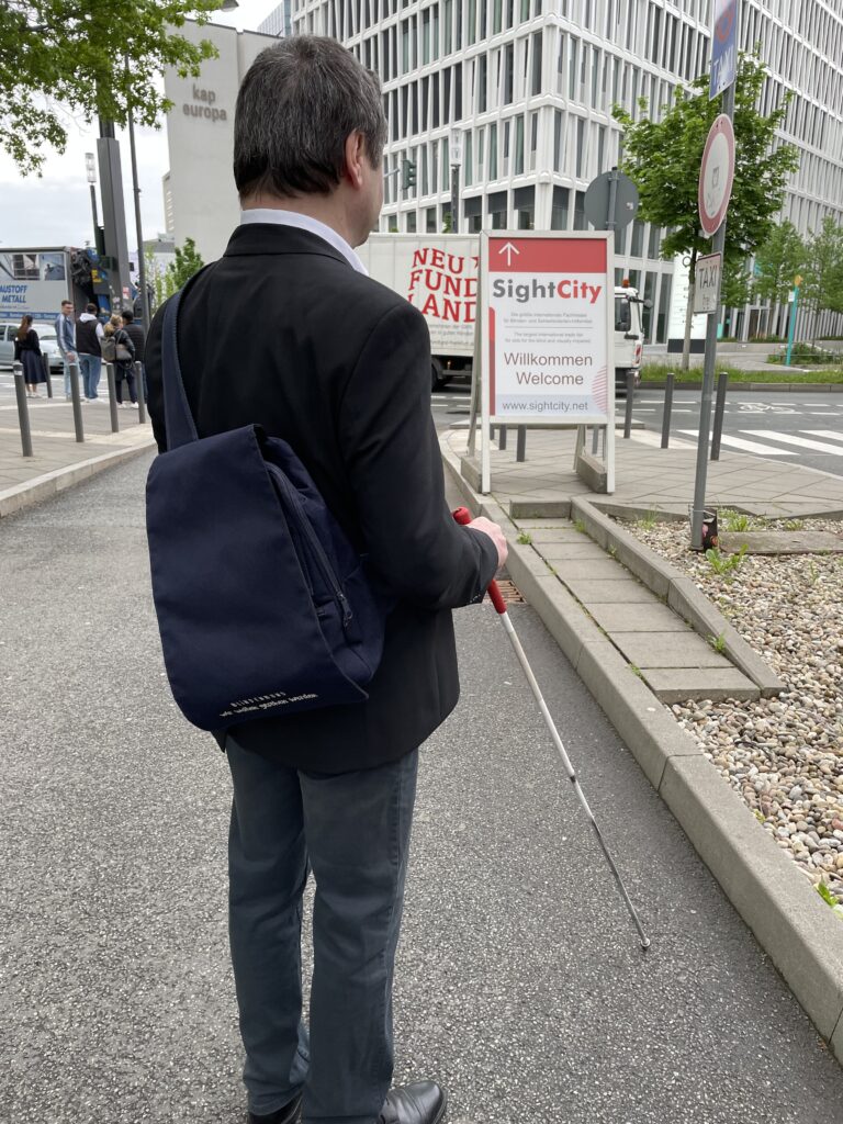 Rear view of a person in a dark blazer with a dark blue backpack bearing the text 'Blindenbund – wir wollen gesehen werden' (Blind Association – we want to be seen). The person holds a white cane with a red handle and stands in front of a SightCity directional sign. The sign shows an upward arrow and the text: 'SightCity – The largest international trade fair for aids for the blind and visually impaired – Willkommen / Welcome – www.sightcity.net'. In the background, the inscription 'kap europa' is visible on a building.