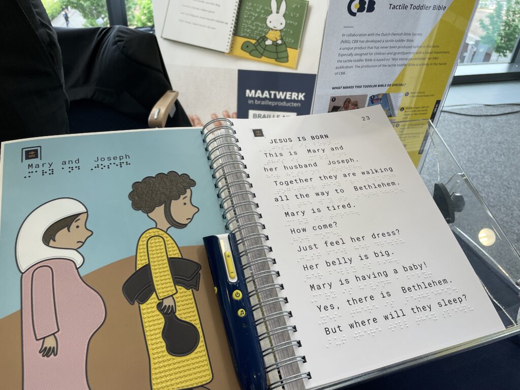 An open book on a table: the 'Tactile Toddler Bible' by publisher CBB. The left page shows a colourful illustration of Mary and Joseph with tactile elements and Braille text. The right page contains the large-print English text of the story 'Jesus is Born' along with Braille lines. A blue audio pen lies between the pages. In the background, information materials and a banner reading 'MAATWERK in brailleproducten' and the CBB logo.