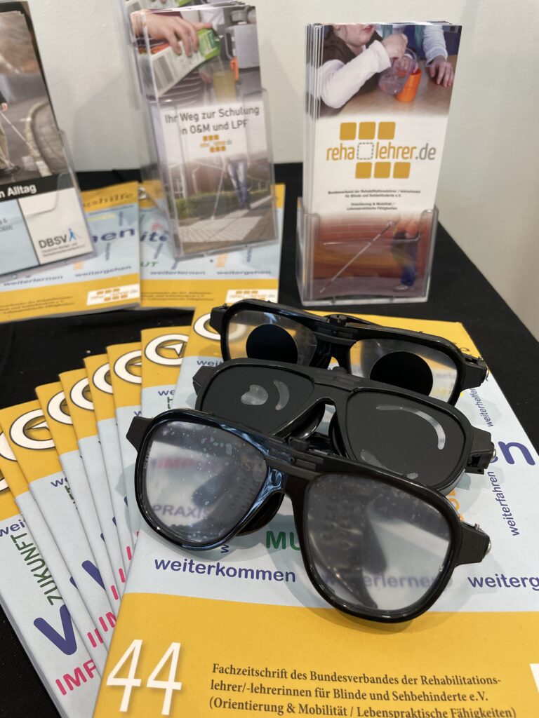 Exhibition table with information materials from the Federal Association of Rehabilitation Teachers for the Blind and Visually Impaired (reha-lehrer.de). On the table are several copies of the specialist journal 'IMF 44' and brochures on orientation & mobility and daily living skills. In the foreground, two black simulation glasses for demonstrating various visual impairments. In the background, brochure holders with additional information materials, including from the DBSV.