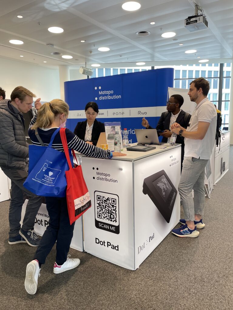 Busy exhibition stand of Matapo Distribution at SightCity. The white counter displays a product image of the 'Dot Pad' and a QR code labelled 'Scan Me'. Behind the counter stand three staff members, including a woman with a laptop. Visitors stand in front of the booth; one carries a blue ViewPlus bag, another a red SightCity bag. In the background, the blue stand backdrop system with logos of Matapo Distribution, dot, eone, and Rivo.