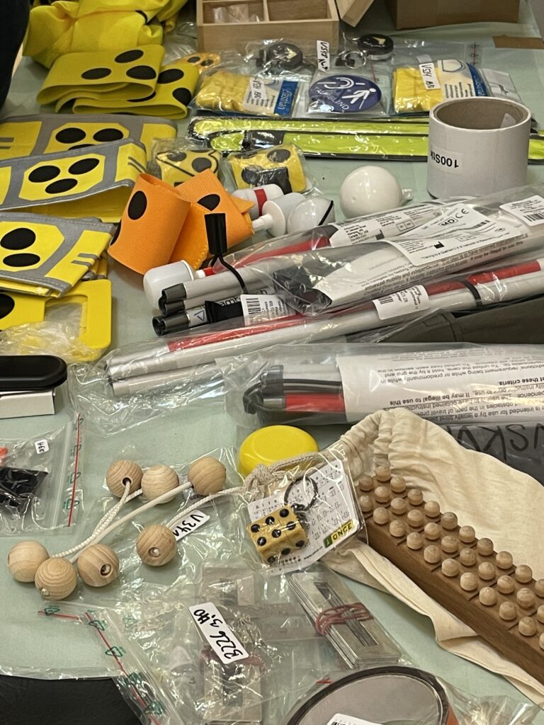 Top-down view of a table covered with a variety of assistive devices for blind and visually impaired people. Visible items include yellow tactile floor markers with black dots, orange and yellow cane grip pieces, several folded white canes in packaging, wooden beads on a cord, a tactile wooden board with knobs, a yellow cap, a white cup, and various small items and accessories in plastic bags. Items are labelled with price tags.