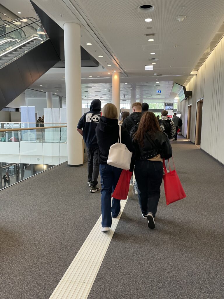 Several people are walking away from the camera through a bright, modern event corridor. In the foreground, two women are visible, one carrying a white bag with red accents, the other holding a red tote bag. A tactile guiding strip runs along the floor. An escalator is visible on the left, and the corridor opens into a larger area in the background. A backpack bears the text "Nature Needs Heroes.
