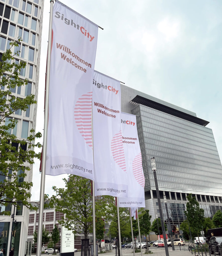 Three white flags bearing the SightCity logo and the bilingual text "Willkommen / Welcome" as well as the web address www.sightcity.net are flying in front of a modern high-rise complex made of glass and concrete. Green trees are visible on the left side of the image. The sky is slightly overcast.