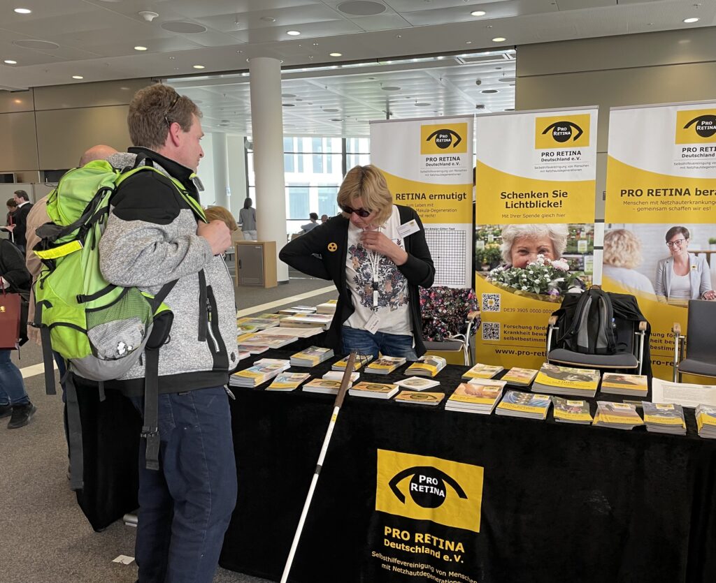 A man with a green-black backpack and a white cane is standing at the PRO RETINA Deutschland e.V. booth at SightCity. Behind the black-covered table, which is laid out with numerous yellow-and-black informational brochures, a woman wearing dark sunglasses is speaking with the visitor. In the background, several PRO RETINA roll-up banners with the slogans "Schenken Sie Lichtblicke!" (Give Moments of Hope!) and "PRO RETINA berät" (PRO RETINA advises) are visible.