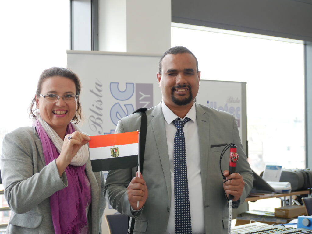 Two people are standing smiling in front of a booth at SightCity. The woman on the left is wearing glasses, a grey blazer, and a purple-and-white scarf. The man on the right is wearing a grey suit with a tie and is holding a folding white cane and an Egyptian flag. The woman is also holding a small Egyptian flag. In the background, a roll-up banner with the text "Blista" is visible.