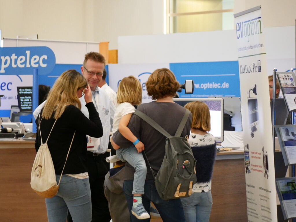Mehrere Personen stehen am Stand von Optelec auf der SightCity. Im Vordergrund sind zwei Frauen von hinten zu sehen, eine davon trägt ein Kleinkind auf dem Arm. Ein Mann mit Brille und weißem Hemd, vermutlich ein Standmitarbeiter, spricht lächelnd mit den Besucherinnen. Im Hintergrund sind Ausstellungswände mit dem Optelec-Logo und der Webadresse www.optelec.de sowie ein Roll-up-Banner von evoptron mit dem Produkt „DaVinci Pro" zu sehen.