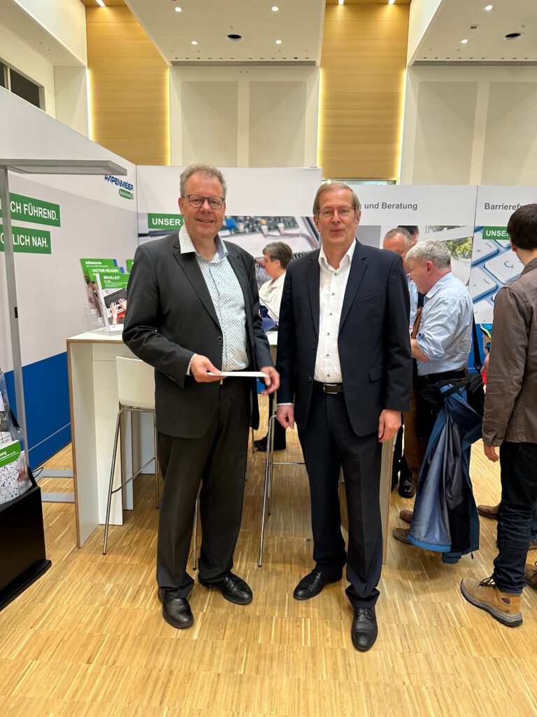 Two men in suits are standing side by side at the Papenmeier RehaTechnik booth at SightCity Frankfurt. The man on the left is holding a white tablet. In the background, exhibition panels with the slogans "Technisch führend. Persönlich nah." and product references to Milestone, Victor Reader, and BRAILLEX are visible.