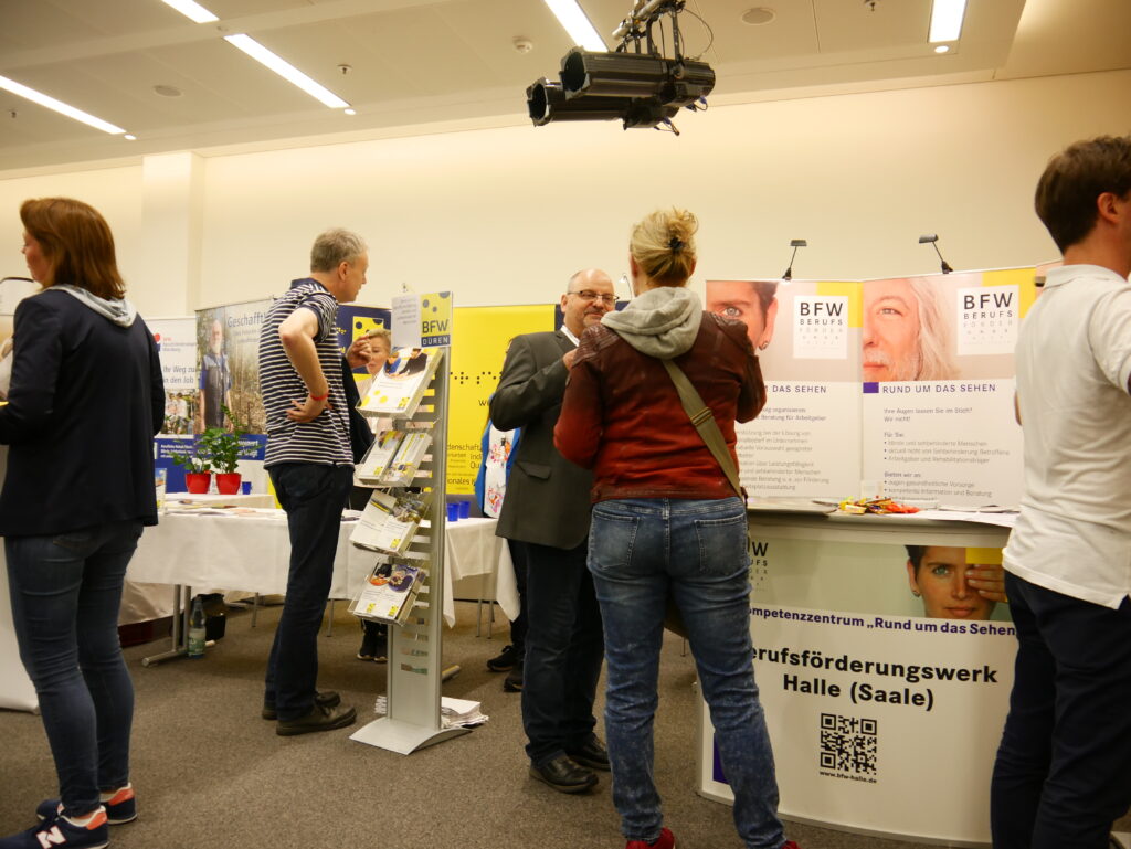 A view of several exhibition booths of vocational rehabilitation centers (BFW) at SightCity. Several visitors and booth staff are engaged in conversation. A brochure stand is visible in the foreground. In the background, banners from BFW Düren and the Berufsförderungswerk Halle (Saale), focusing on the "Competence Center for Vision," are visible. A QR code is displayed on the booth banner.