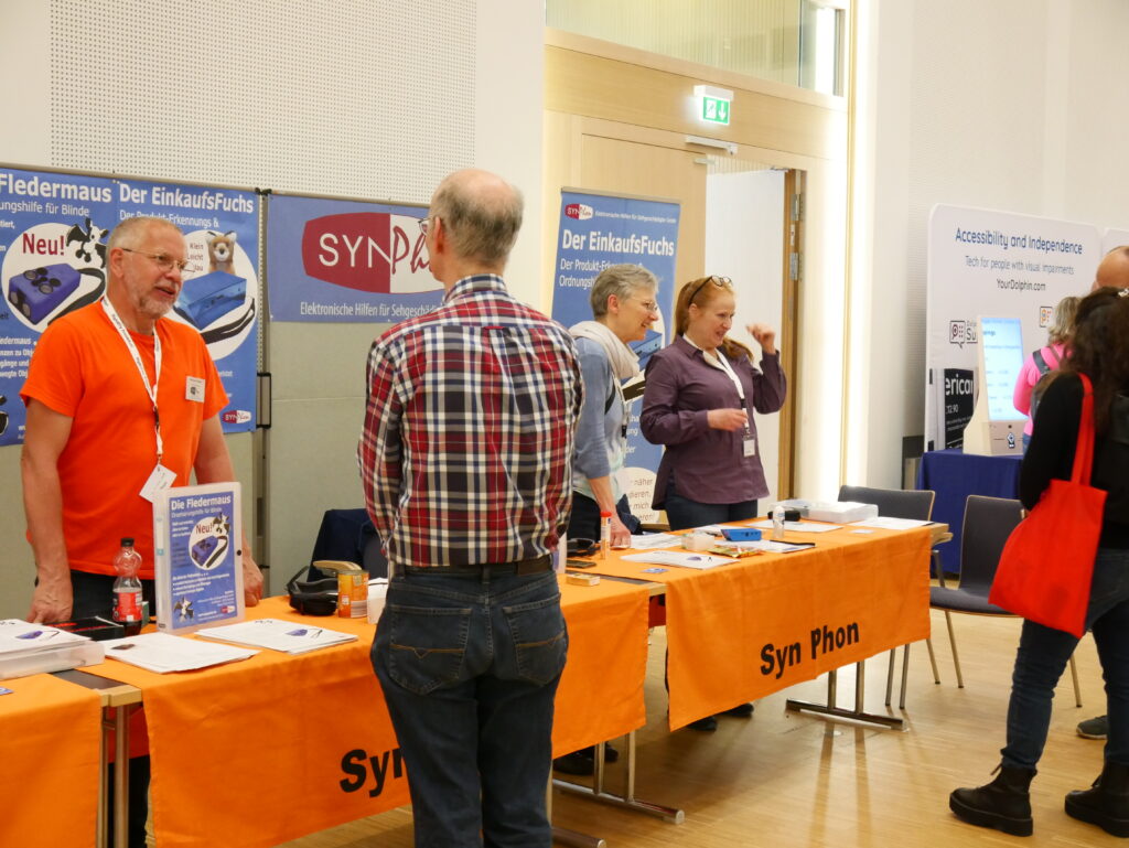 A view of the SynPhon booth at SightCity Frankfurt. A man in an orange T-shirt stands on the left side of the booth, talking to a visitor. On the orange table, informational materials and products are displayed, including "Die Fledermaus" (an orientation aid for blind people) and "Der EinkaufsFuchs." In the background, additional exhibition booths, including one with the YourDolphin logo, are visible.