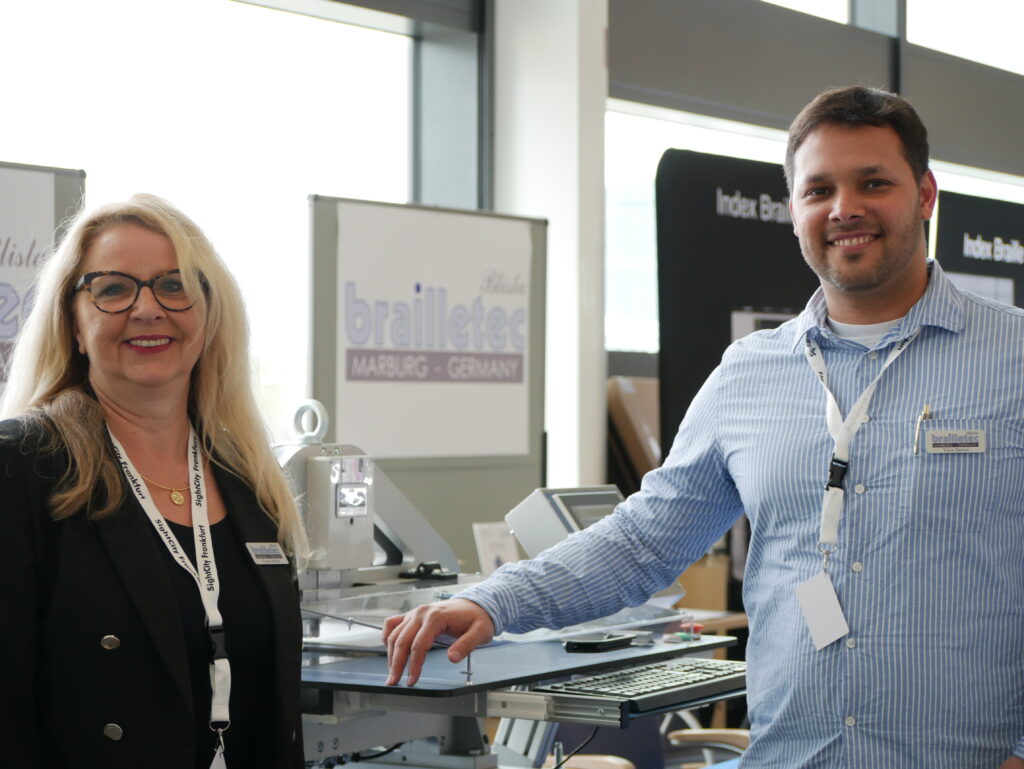 A woman with blonde hair and glasses and a man in a light blue shirt are standing smiling at the brailletec (Marburg, Germany) booth at SightCity Frankfurt. In front of them is a Braille embosser on a table. In the background, additional exhibition areas with the label "Index Braille" are visible.
