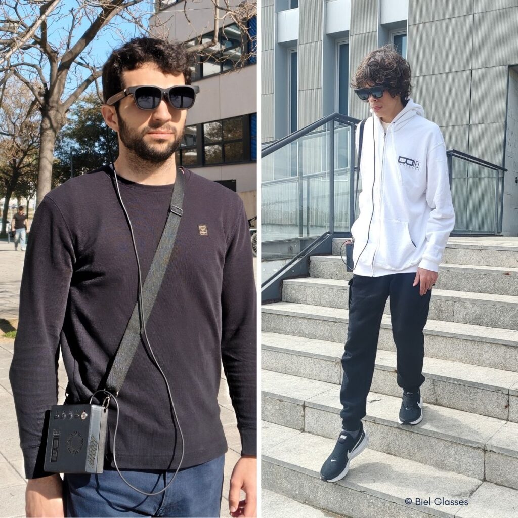 Zwei Männer tragen Biel Glasses im Freien: links ein Mann mit dunklem Pullover und umgehängtem Gerät, rechts ein junger Mann in weißem Hoodie auf einer Treppe vor einem Gebäude.