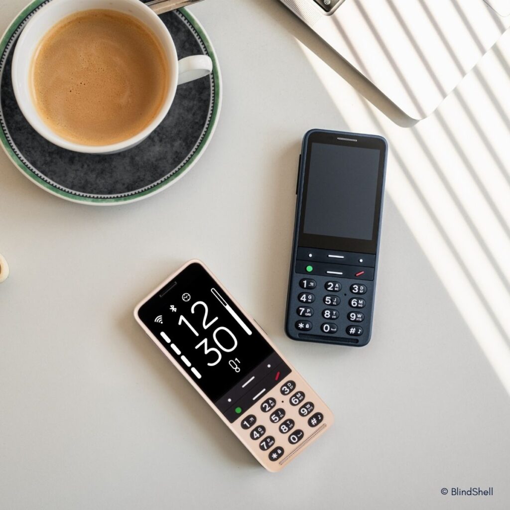 Zwei BlindShell-Mobiltelefone mit großen Tasten auf einem Tisch: eines in Rosa mit Display (12:30 Uhr), eines in Dunkelblau. Daneben eine Kaffeetasse.