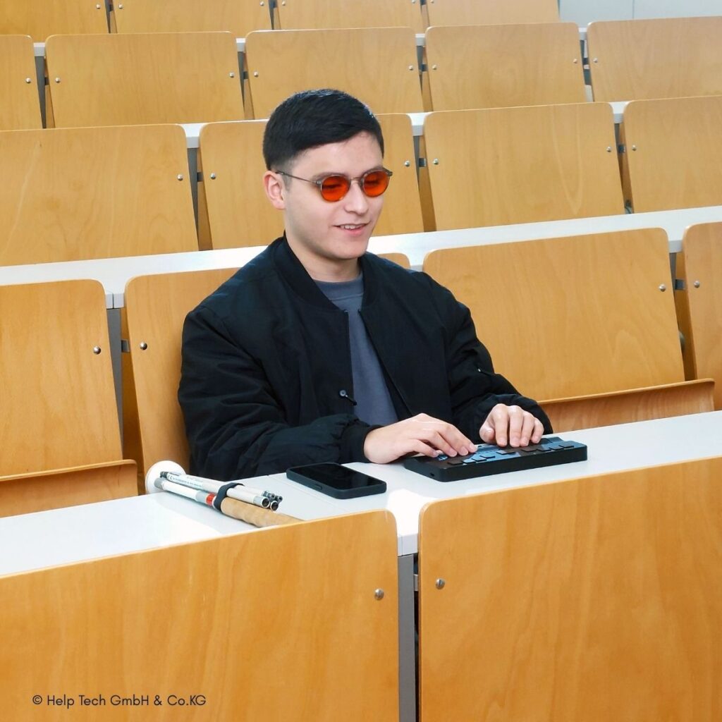 Junger Mann mit orangefarbener Tönung-Brille liest an einem Hörsaaltisch eine Braillezeile von Help Tech. Neben ihm liegen Smartphone und zusammengeklappter Langstock.
