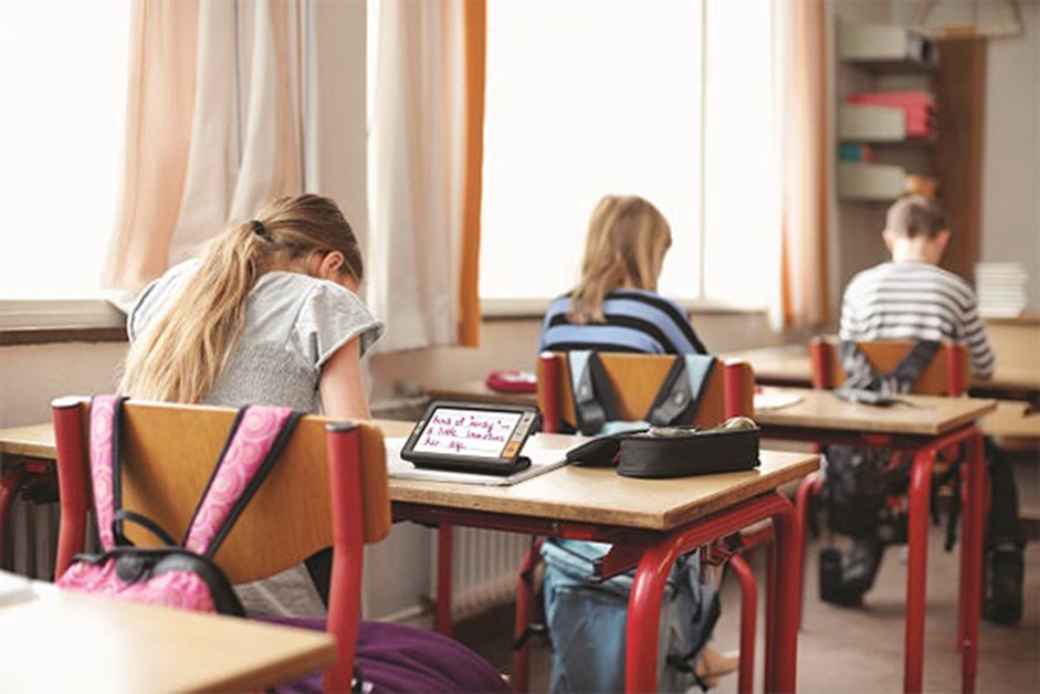 A schoolgirl is sitting at a desk in a classroom, using an electronic magnifying glass on the table next to her.