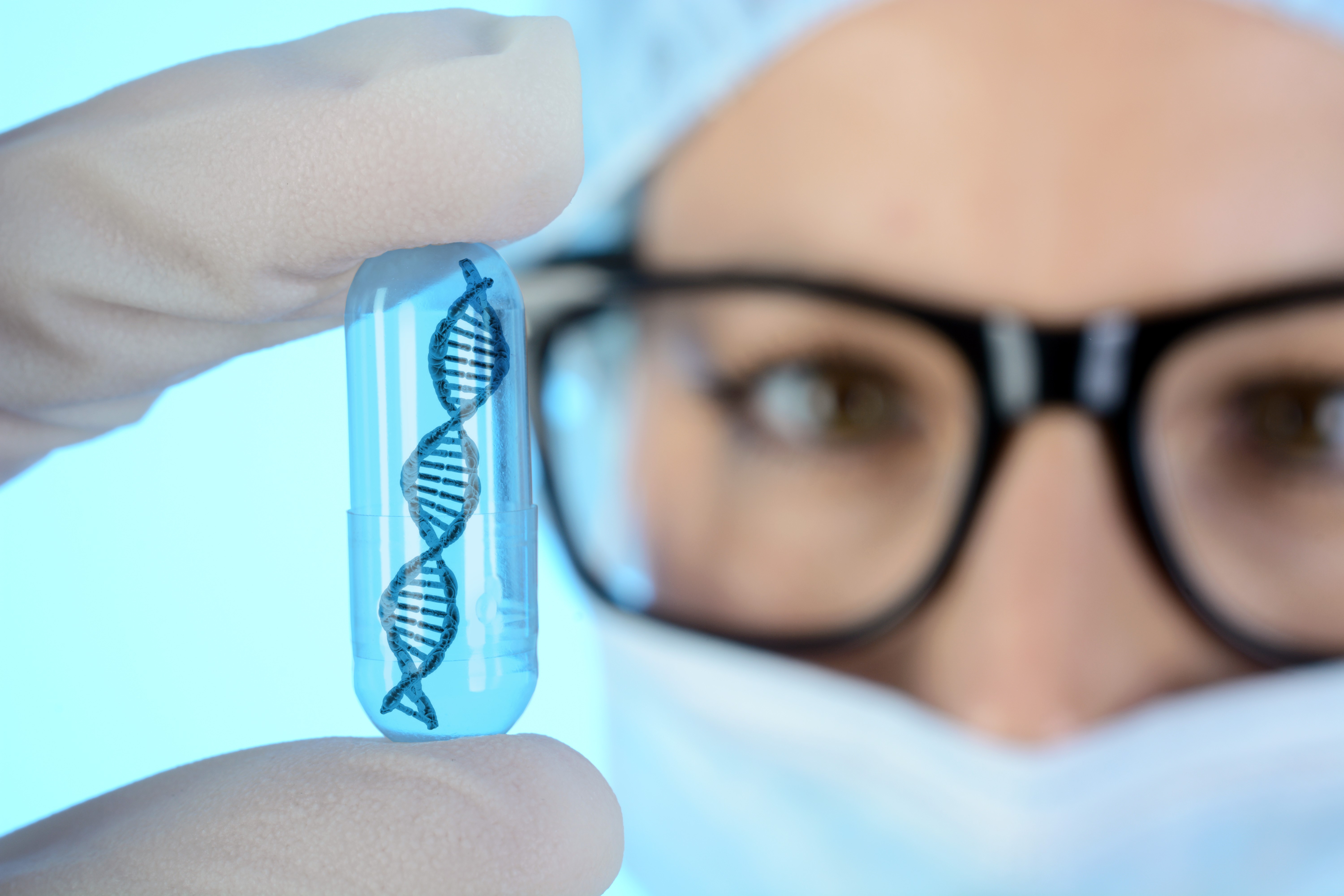A person is holding a double helix inside a transparent pill between two fingers. They are wearing glasses, a face mask, and a cap.